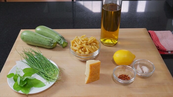 ingredients for the pasta d recipe displayed on the chopping board. lemon, garlic, olive oil, parmesan, herbs, courgette, pasta, dried chilli,