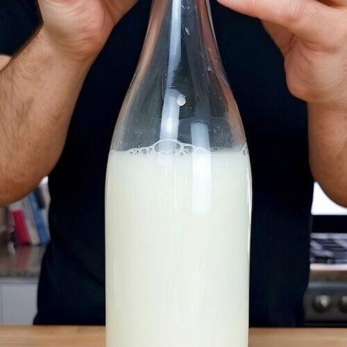 A person sealing a glass bottle filled with creamy homemade oat milk on a kitchen countertop.