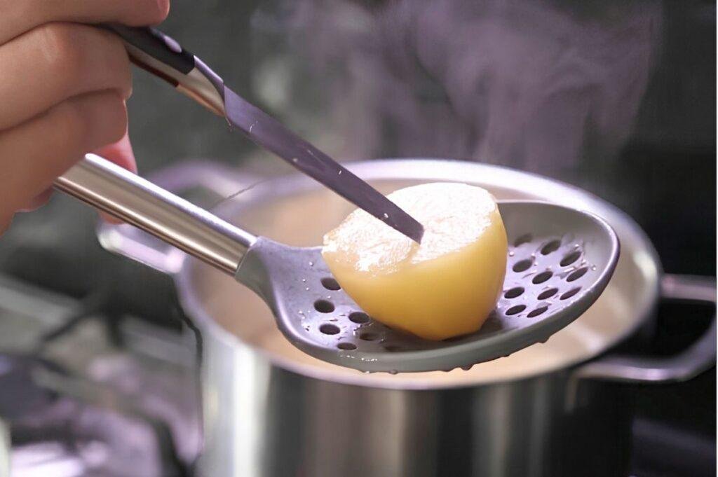 Slotted spoon lifting a parboiled potato from a pot of steaming water