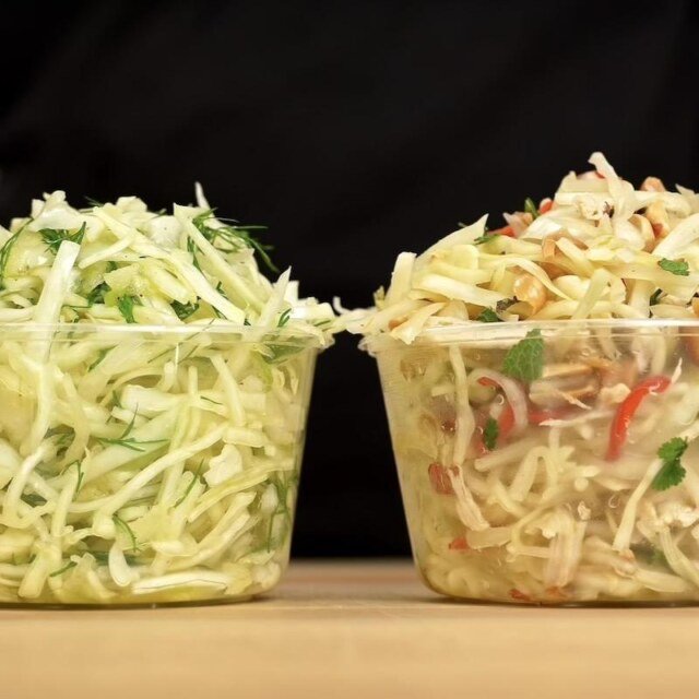 2 containers filled abundantly with different fresh slaws on a wooden counter top