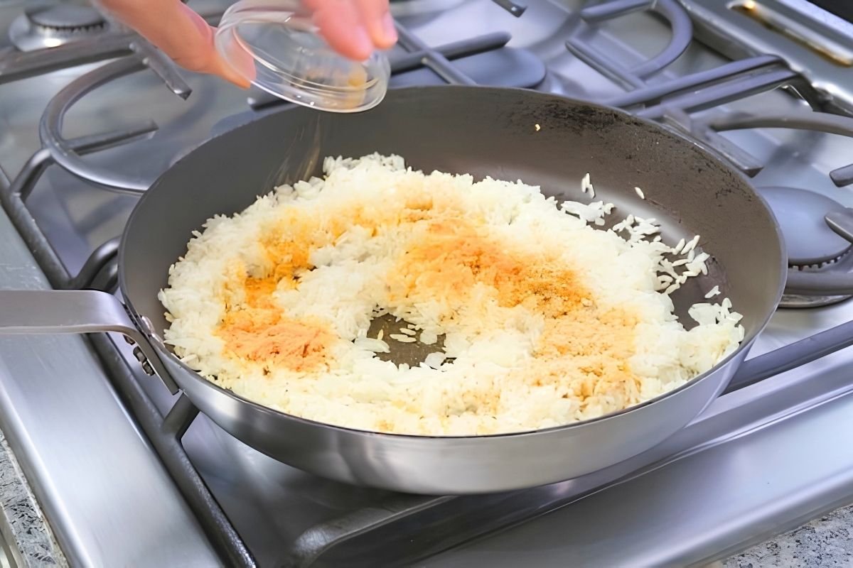 Hand tipping a glass dish of seasoning over rice thats frying in a pan