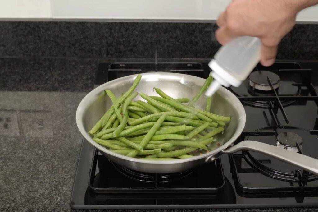 Sauce being squeezed from a bottle over green beans in a steel pan on the hob