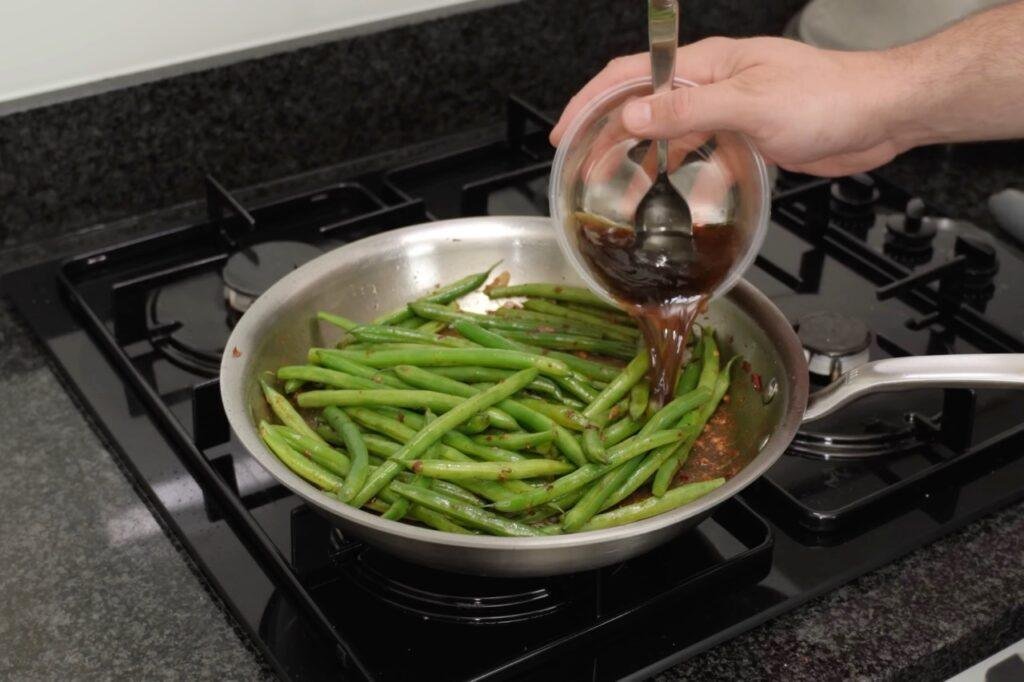 Spoon pouring dark sauce over green beans in a steel pan on a gas hob