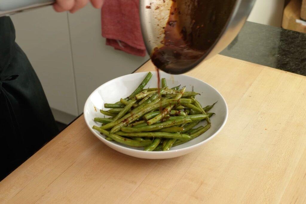 Glossy dark sauce being poured from a pan over green beans in a white bowl