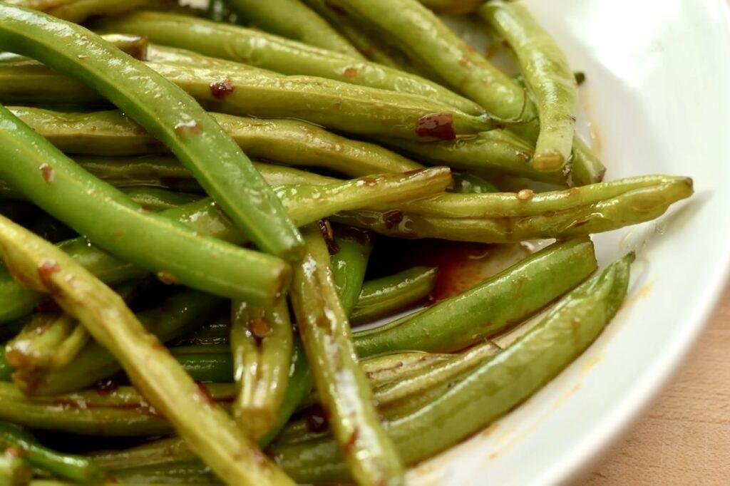 Close up of charred green beans coated in dark sauce in a white bowl