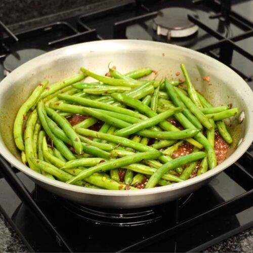 Steel pan full of green beans in dark sauce with chilli on a gas hob