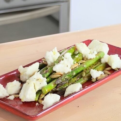 Red dish of sauteed asparagus topped with crumbled feta and toasted pumpkin seeds