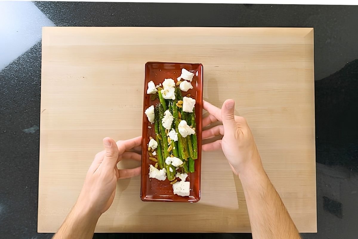 Overhead view of hands holding a red dish of sauteed asparagus topped with crumbled feta