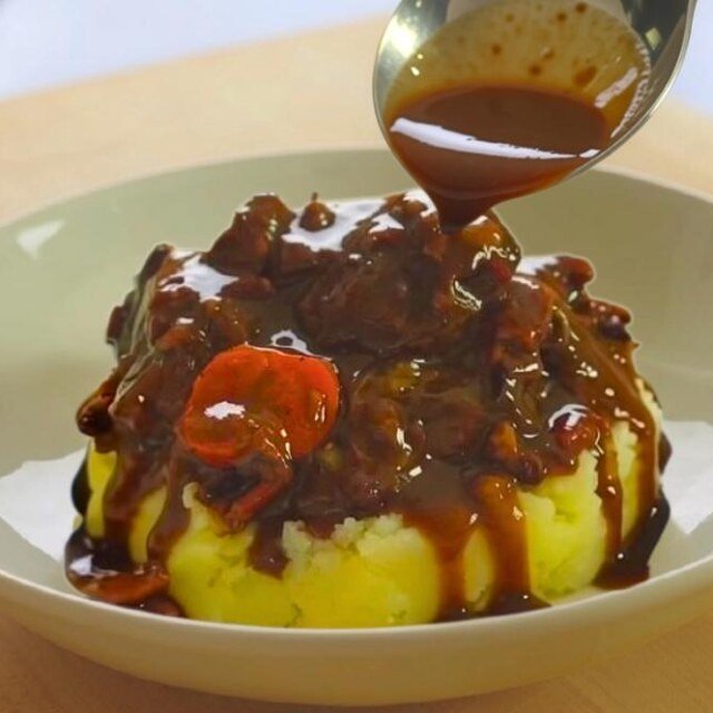 Spoon pouring rich red wine sauce over beef bourguignon on mashed potato in a white bowl