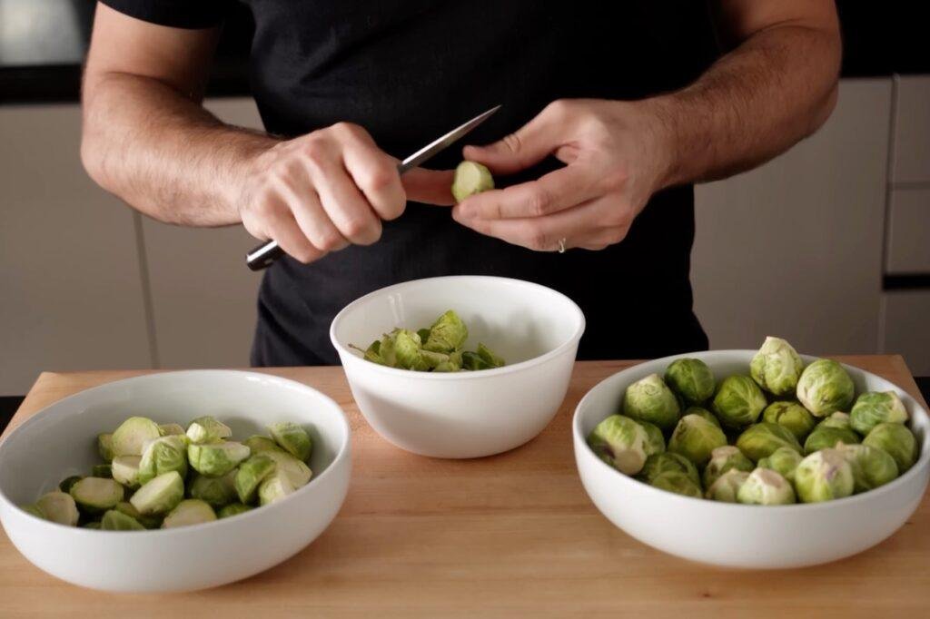 Person halving Brussels sprouts with a small knife beside bowls of whole and halved sprouts