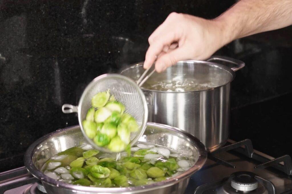 Strainer lowering blanched Brussels sprout halves into a bowl of iced water beside a pot