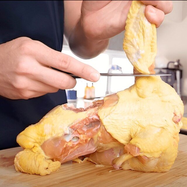 Chef using a knife to butcher a whole raw chicken on a wooden board