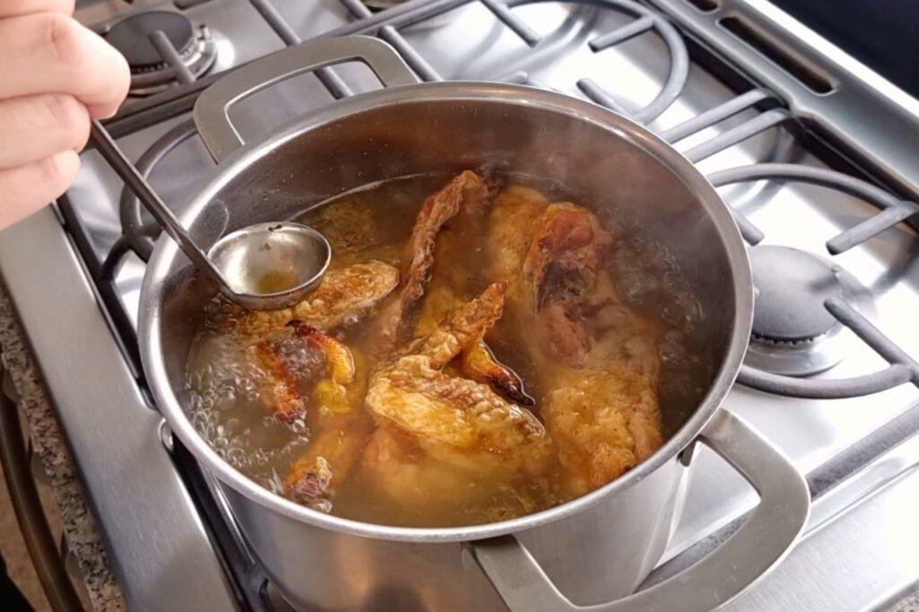 Browned chicken pieces in broth in a steel pot with a ladle