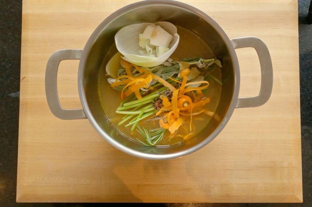 Overhead view of a pot of clear broth with carrot, celery and herbs on a wooden board