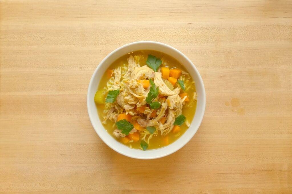 Overhead view of a white bowl of chicken noodle soup with carrot and herbs on a wooden counter