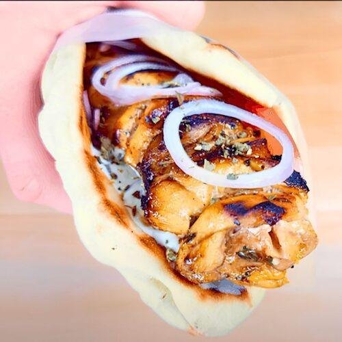 Hand holding a chicken souvlaki pitta wrap with onion rings and tzatziki