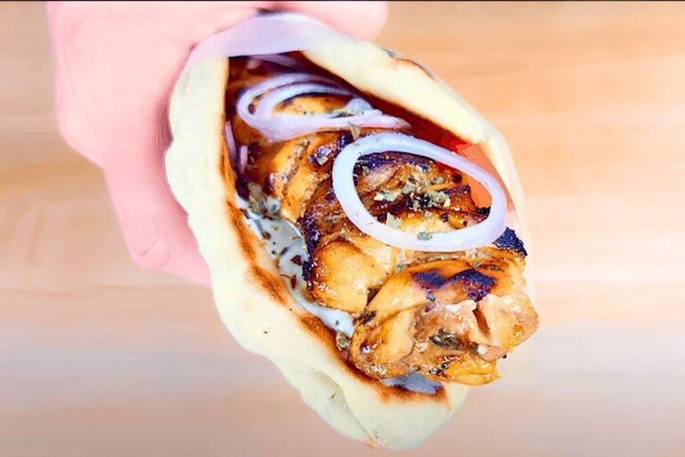 Hand holding a chicken souvlaki pitta wrap with onion rings and tzatziki