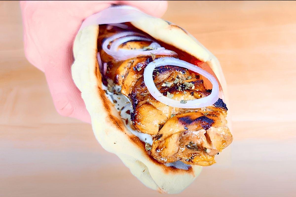 Hand holding a chicken souvlaki pitta wrap with onion rings and tzatziki