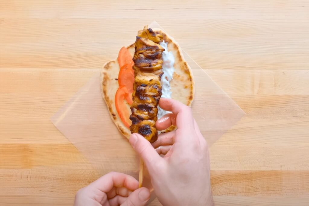 Hand pulling a chicken skewer out of a pitta with tomato and tzatziki