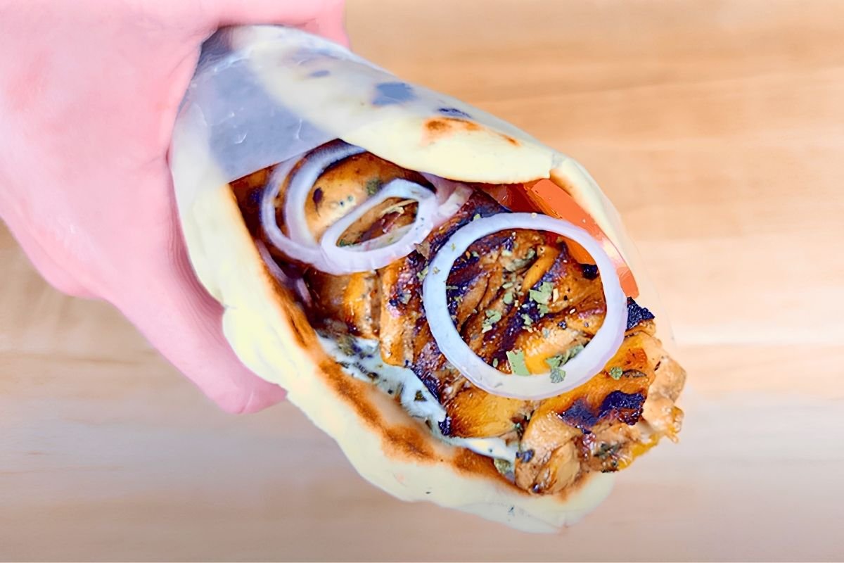 Hand holding a chicken souvlaki pitta wrapped in paper with onion rings and tomato