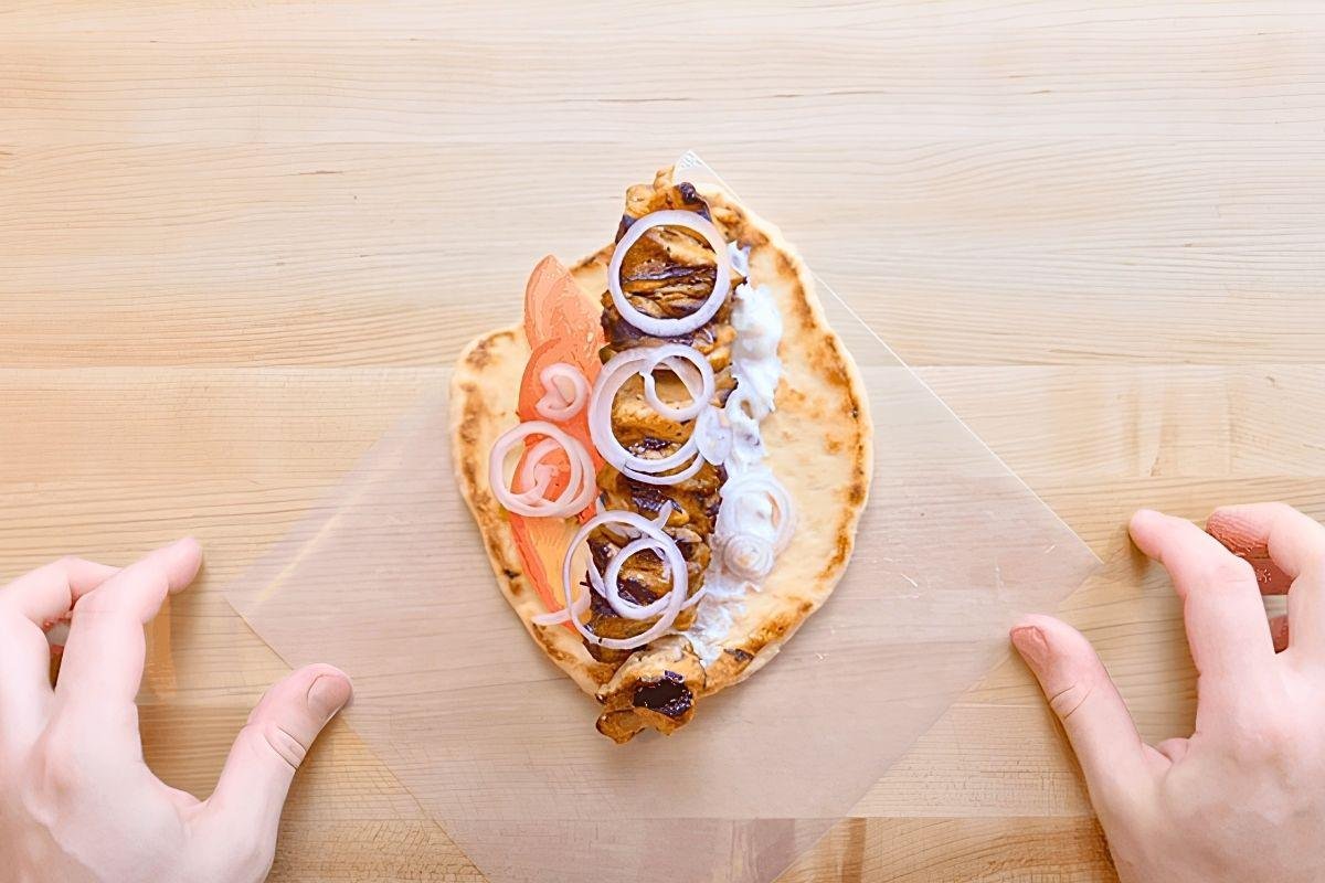 Overhead view of a pitta with a chicken skewer, tomato, onion rings and tzatziki on greaseproof paper