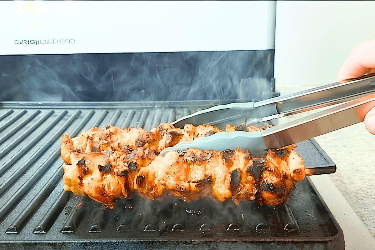Tongs turning a chicken souvlaki skewer on a smoking grill pan