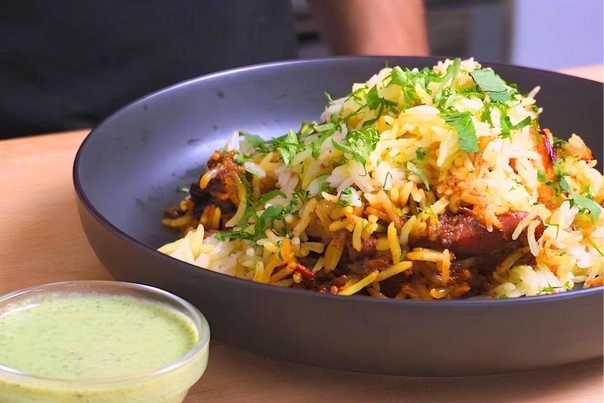 Dark bowl of chicken biryani topped with fresh herbs, with a small glass bowl of green raita beside it
