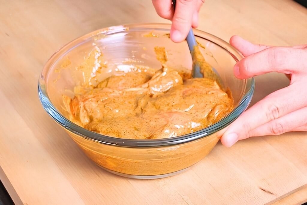 Chicken being mixed into a spiced yoghurt marinade in a glass bowl