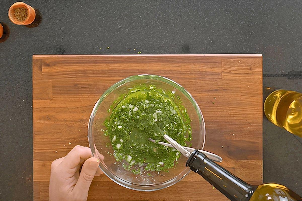 Overhead view of chimichurri being mixed in a glass bowl while olive oil is poured in