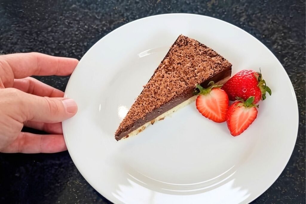 Hand holding a white plate with a slice of chocolate tart, grated chocolate and fresh strawberries