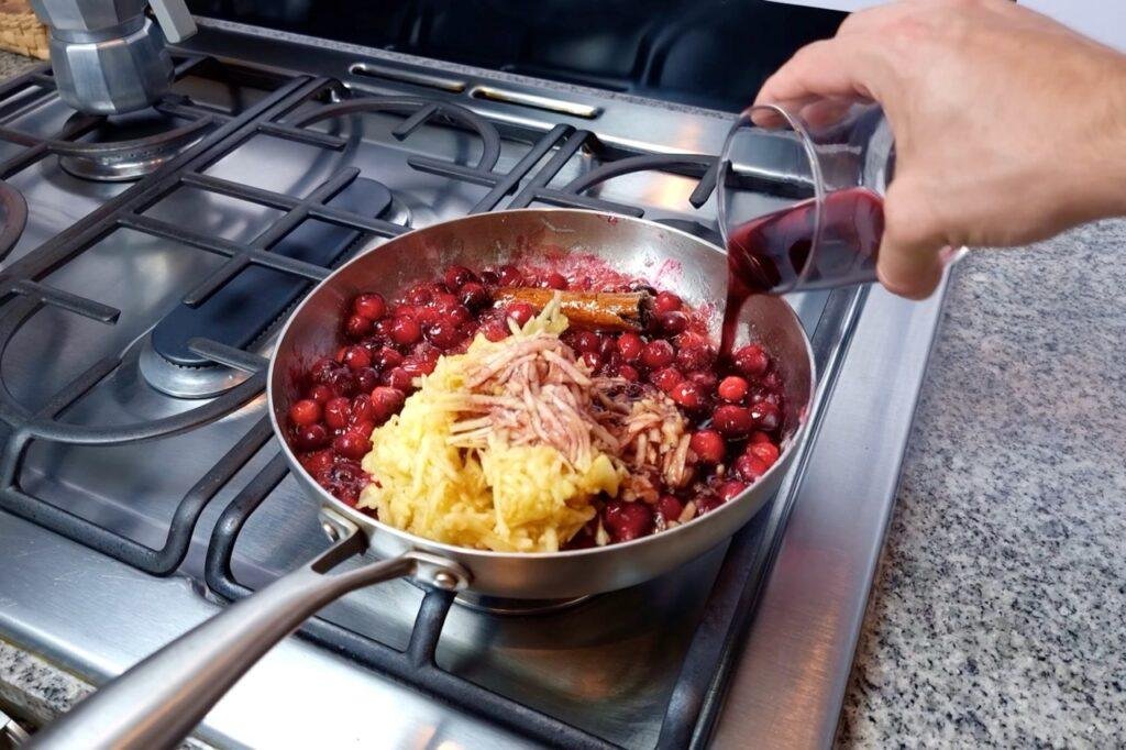 Grated ginger on top of cranberries simmering in a steel pan, with a hand pouring in liquid