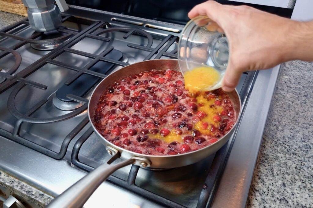 Hand pouring orange juice into a pan of cranberries on the hob