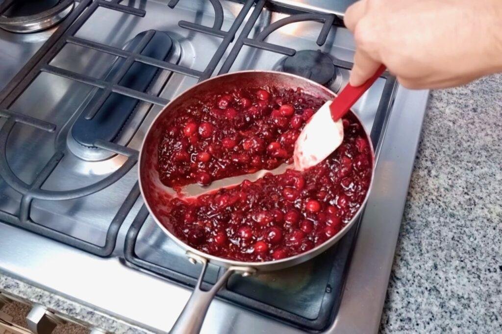 Hand stirring thickened cranberry sauce in a steel pan with a red spatula