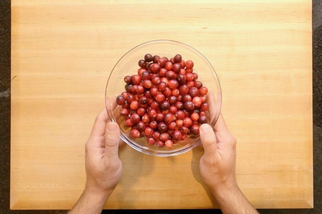 Overhead view of hands holding a glass bowl of fresh cranberries on a wooden board