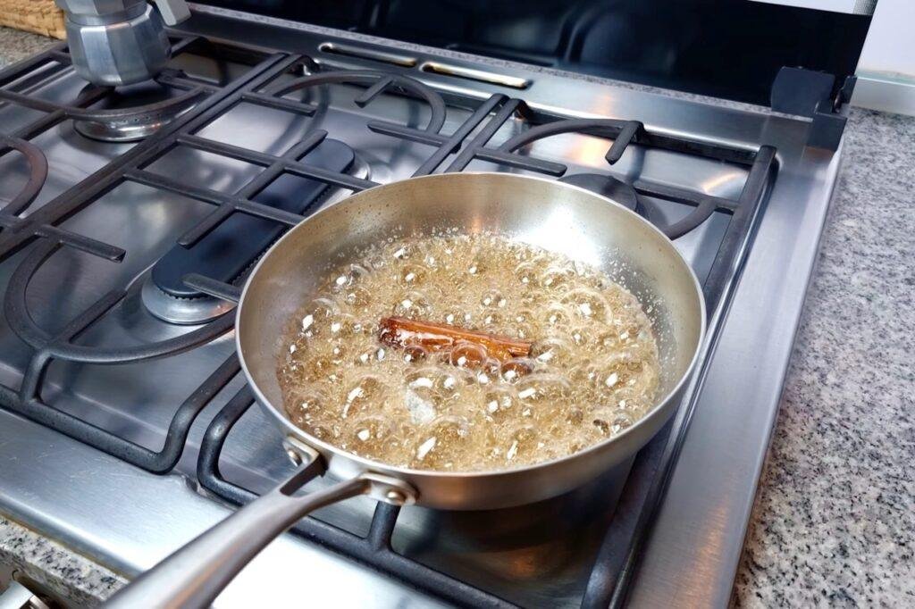 Sugar syrup bubbling with a cinnamon stick in a steel pan on the hob