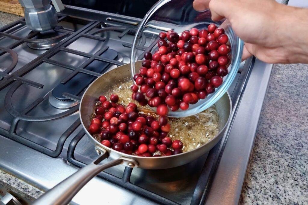 Fresh cranberries being tipped from a glass bowl into a steel pan on the hob