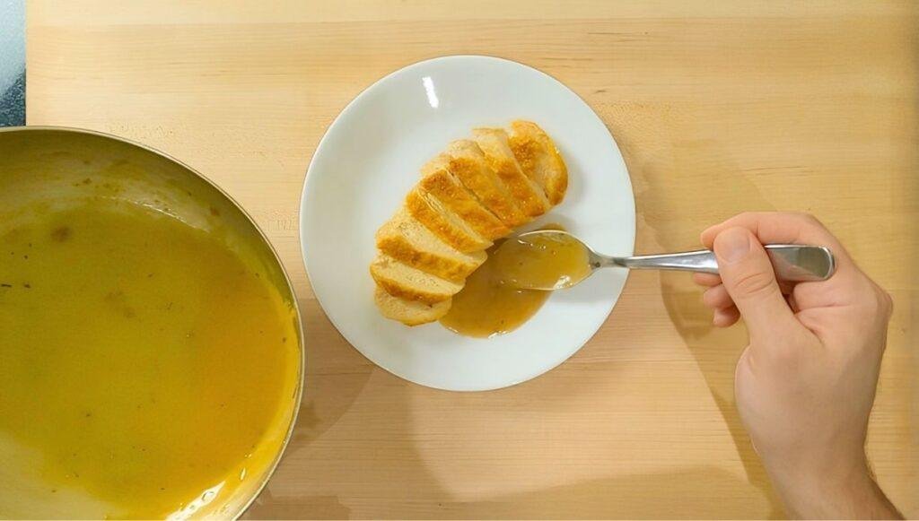 Overhead view of sliced chicken breast on a white plate being spooned with golden pan sauce, beside the pan