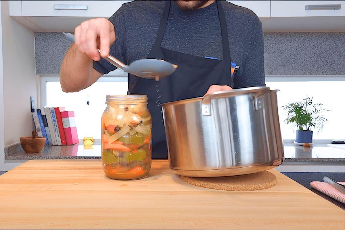 Chef spooning hot pickling liquid into a jar of chillies and vegetables beside a steel pot