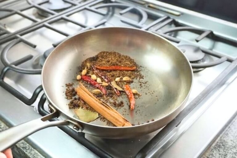 Steel frying pan of toasting whole spices with cinnamon, dried chillies and a bay leaf on a gas hob