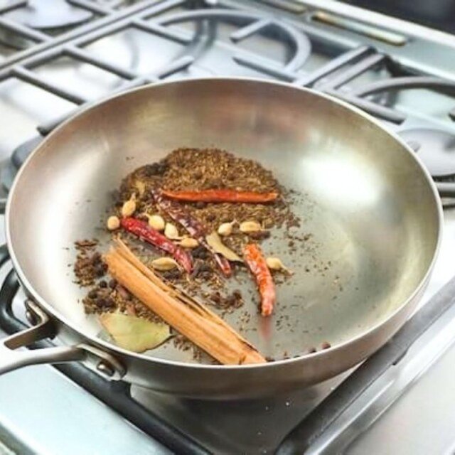 Steel frying pan of toasting whole spices with cinnamon, dried chillies and a bay leaf on a gas hob