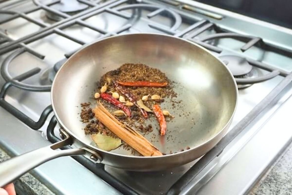 Steel frying pan of toasting whole spices with cinnamon, dried chillies and a bay leaf on a gas hob