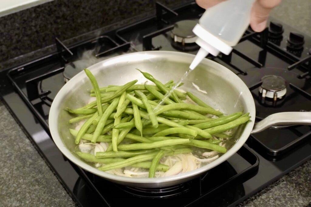 Water being squeezed from a bottle over green beans in a steel pan