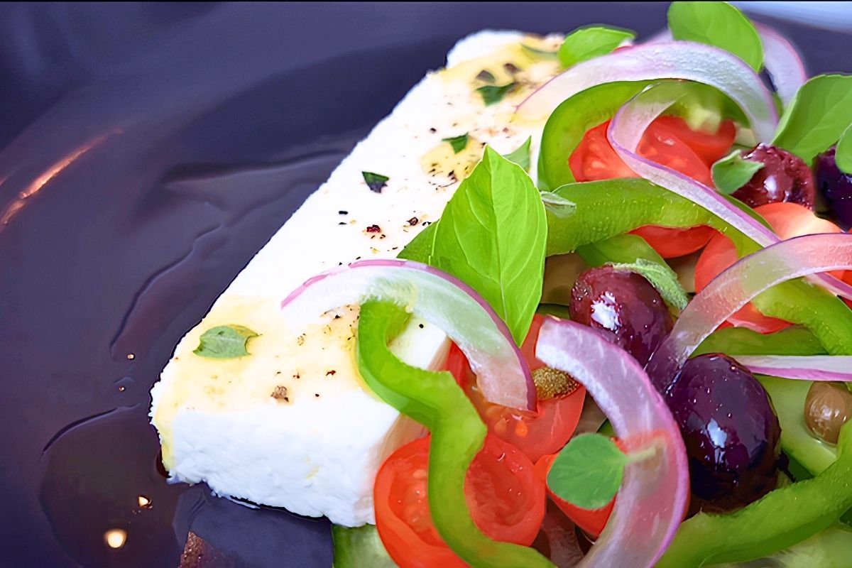 Close up of feta topped with oregano and olive oil beside green pepper, red onion, tomatoes and olives