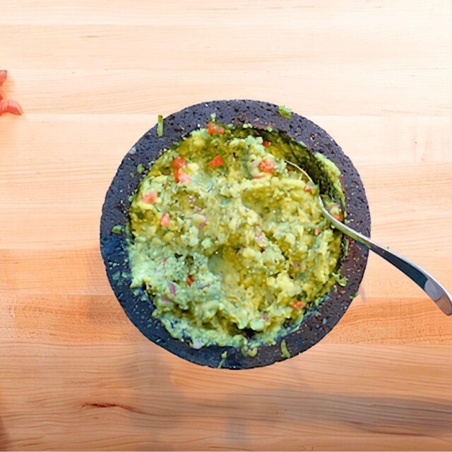 Overhead view of guacamole in a molcajete with a spoon and a pile of diced tomato on a wooden board