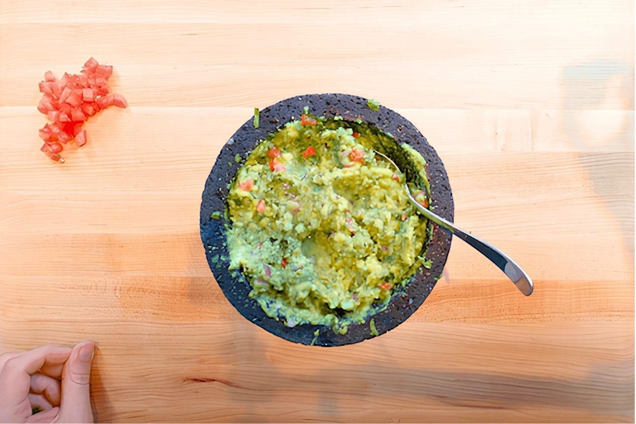 Overhead view of guacamole in a molcajete with a spoon and a pile of diced tomato on a wooden board