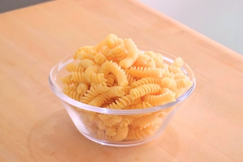 Glass bowl of dry fusilli pasta on a wooden board
