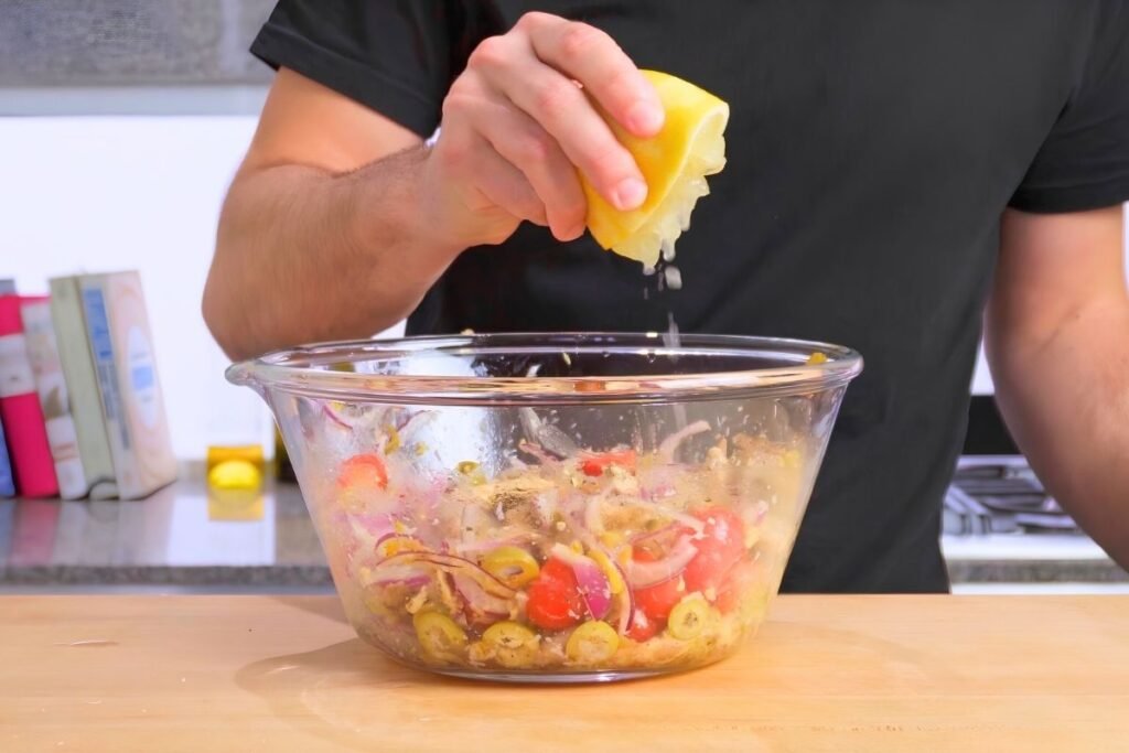 Hands squeezing lemon over a glass bowl of pasta salad with onion, olives and tomatoes