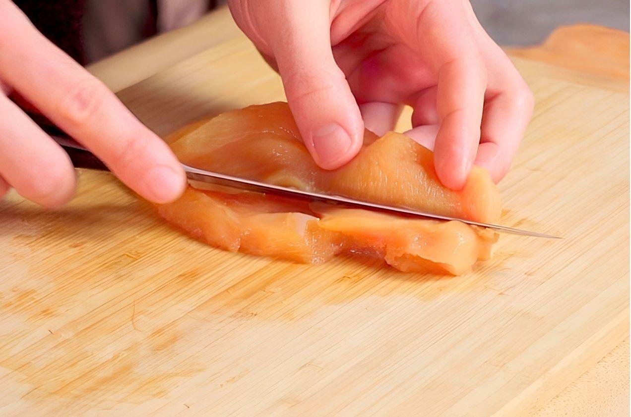 Hands slicing a chicken breast in half horizontally with a knife on a board