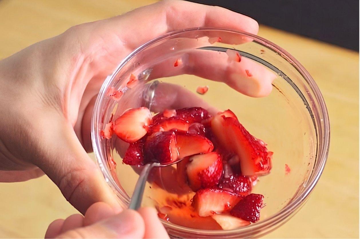 Hand holding a glass bowl of sliced strawberries in juice with a spoon