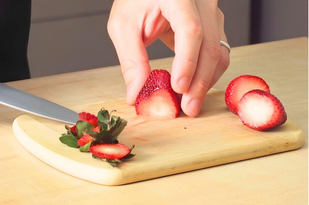 Hands slicing a strawberry on a small wooden board with a knife
